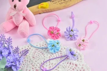 Пять заколок для волос с цветами из ткани лежат на белой ажурной салфетке рядом с мягкой розовой игрушкой-медведем и веточкой лаванды.