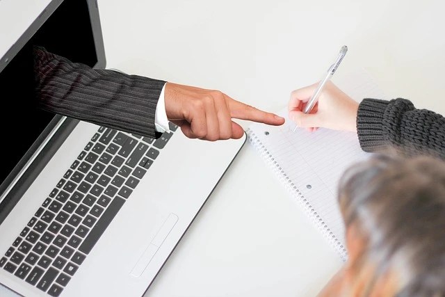 Рука в деловом костюме, выходящая из экрана ноутбука, указывает пальцем на тетрадь, в которой человек делает записи.