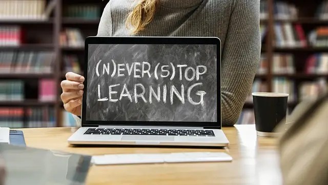 Открытый ноутбук с надписью "(N)EVER (S)TOP LEARNING" (Никогда не прекращай учиться), написанной мелом на черном фоне, которую держит человек в библиотеке или классе.