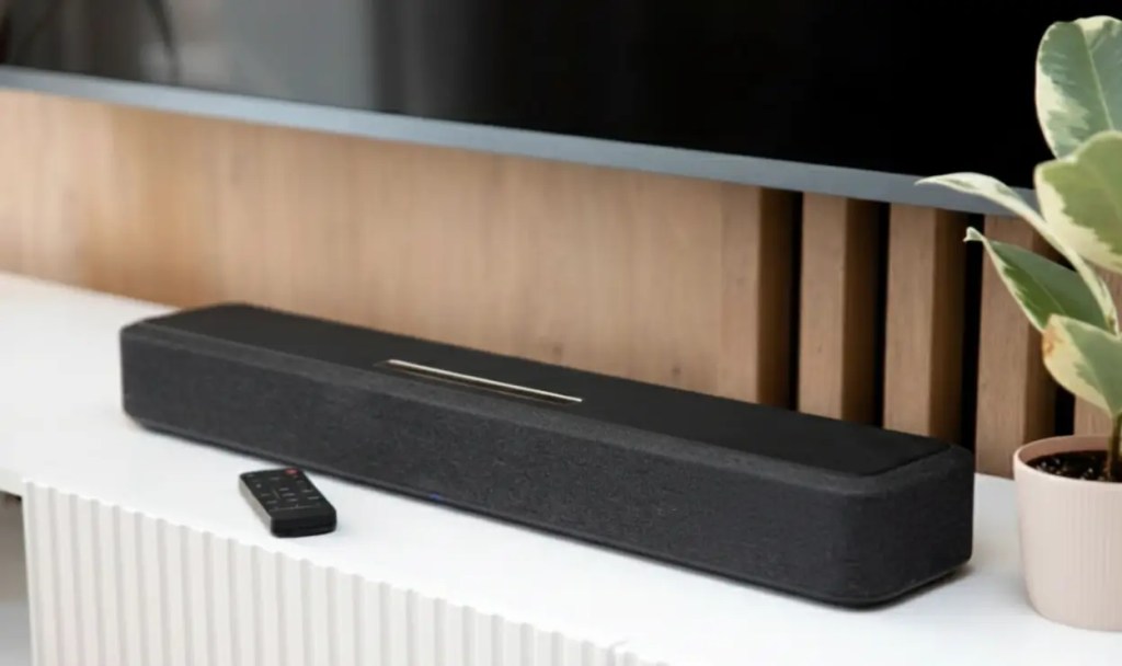 Черный современный саундбар с пультом дистанционного управления на белой тумбе под телевизором в минималистичном интерьере.